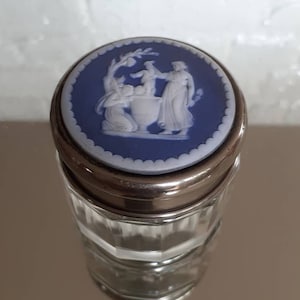 May include: A clear glass jar with a silver rim and a blue lid. The lid features a white relief design of figures. The jar has a faceted design. The jar is sitting on a reflective surface.