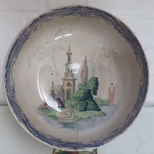May include: A vintage ceramic bowl with a detailed landscape scene in the center. The bowl features a blue and white color scheme with a decorative border. The landscape includes a building, trees, and water, creating a classic aesthetic.