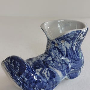 May include: A ceramic boot-shaped vase with a blue and white pattern. The vase has a rounded toe, a high ankle, and an open top. The blue and white design resembles a watercolor painting.