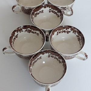 Könnte beinhalten: Sechs cremefarbene Teetassen mit braunen floralen und architektonischen Mustern. Jede Tasse hat einen Henkel und einen dekorativen Rand. Die Tassen sind kreisförmig angeordnet und präsentieren ihr Design.