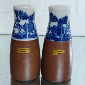 Könnte beinhalten: Ein Paar Salz- und Pfefferstreuer. Die Oberteile sind aus weißem Porzellan mit blauen Landschaftsdesigns. Die Sockel sind aus Holz mit goldfarbenen Buchstaben: "S" und "P". Die Streuer sind etwa 10 cm hoch.