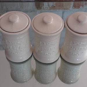 May include: Three off-white ceramic spice jars with lids. Each jar has a curved shape and is labeled with a different spice name: "Garlic", "Proven", and "Basil". The jars are arranged in a row, reflecting on a shiny surface.