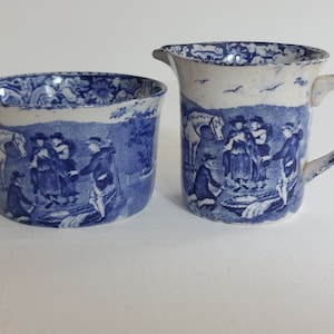 May include: A blue and white ceramic creamer and sugar bowl set. The set features a pastoral scene with figures, horses, and trees. The creamer has a handle and spout. The sugar bowl is round. Both pieces have a white rim.