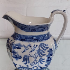 Op de afbeelding: Een wit en blauw keramieken kan met een Willow-patroon. De kan heeft een gebogen handvat en een tuit. Het blauwe ontwerp toont pagodes, vogels en bomen. De kan staat op een reflecterend oppervlak.