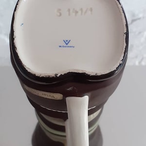 May include: A vintage brown and white ceramic mug with a white handle. The bottom of the mug has the text "S 141/1" and "W. Germany". The mug has a unique shape and design.
