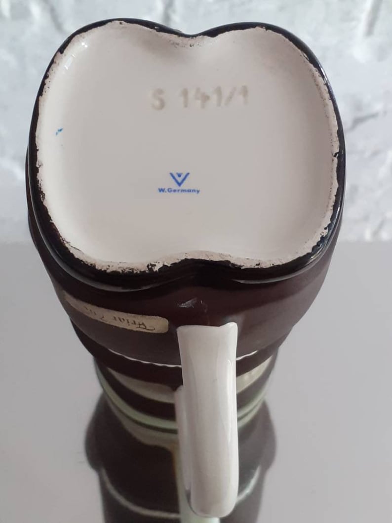 May include: A vintage brown and white ceramic mug with a white handle. The bottom of the mug has the text "S 141/1" and "W. Germany". The mug has a unique shape and design.