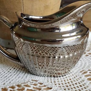 May include: Silver creamer with a handle and spout. The creamer has a textured, patterned design on the lower half and a smooth, reflective surface. It sits on a white, crocheted doily.