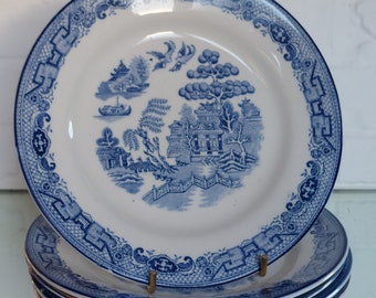 May include: A stack of four white ceramic plates with a blue Willow pattern. Each plate features a detailed landscape scene with pagodas, trees, and a bridge. The plates have a blue border with decorative elements.