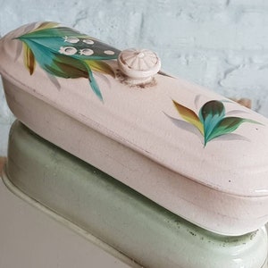 May include: A vintage, oblong, off-white ceramic trinket box with a lid. The lid features hand-painted floral designs in teal, green, and yellow. A small, round knob serves as a handle. The box is reflected in a mirrored surface.