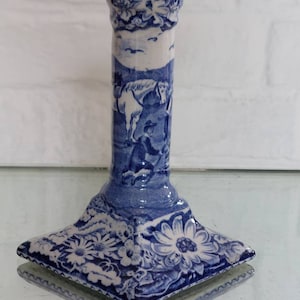 May include: Blue and white ceramic candlestick holder with a square base and floral and pastoral scene designs. The candlestick has a flared base, a cylindrical body, and a circular top. The design features a horse and rider.