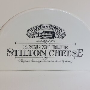 May include: A white, semi-circular cheese container with black text. The text reads "Tuxford & Tebbutt", "English Blue Stilton Cheese", and "Melton Mowbray, Leicestershire, England". The container has a vintage aesthetic.