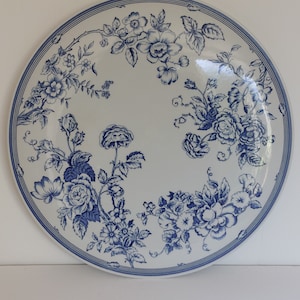 May include: A round, white ceramic plate with a blue floral design. The plate features a detailed pattern of blue flowers and leaves, with a double blue line border. The floral design is arranged around the edge of the plate.
