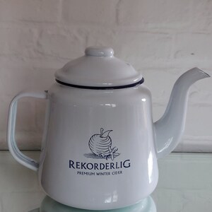 May include: White enamel teapot with a lid and spout. The teapot has a dark blue trim and a handle. The front of the teapot features the text "REKORDERLIG PREMIUM WINTER CIDER" with an apple and cinnamon stick graphic.