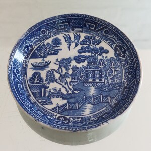 Può includere: Un piatto in ceramica blu e bianco con un motivo Willow. Il piatto presenta una scena paesaggistica dettagliata con un ponte, edifici e alberi. Il bordo ha un bordo decorativo. Il piatto è rotondo e ha un bordo leggermente rialzato.