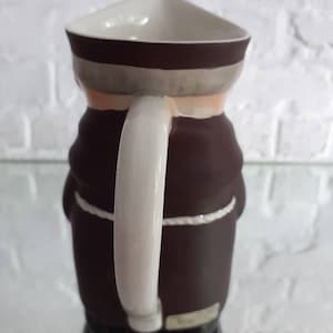 May include: A ceramic pitcher shaped like a monk, featuring a triangular top and a white handle. The pitcher is primarily brown with a white and gray band around the top. A white rope detail accents the waist.