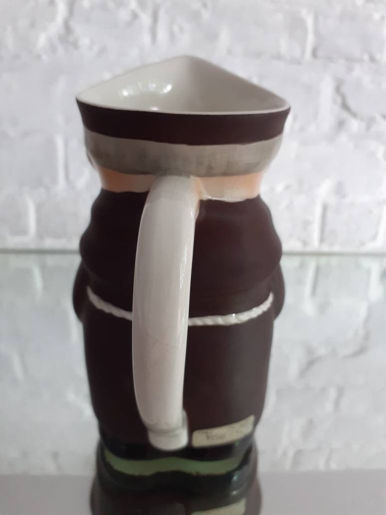 May include: A ceramic pitcher shaped like a monk, featuring a triangular top and a white handle. The pitcher is primarily brown with a white and gray band around the top. A white rope detail accents the waist.