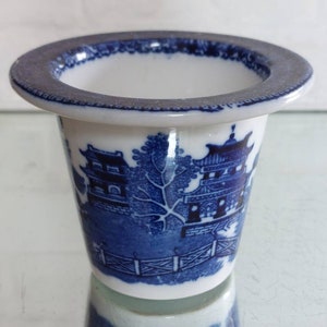 May include: A small, white ceramic container with a blue Willow pattern design. The container has a wide, dark blue rim and a cylindrical shape. The blue pattern depicts a landscape with buildings, trees, and a fence.