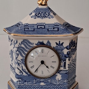 May include: A vintage Wedgwood clock with a blue and white Willow pattern design. The clock has a pagoda-style top with a gold-colored dog figurine. The clock face is round with Roman numerals and the Wedgwood logo. The clock is approximately 6 inches tall.