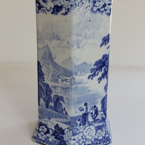 May include: A blue and white ceramic vase with a rectangular shape and a flared base. The vase features a detailed landscape scene with figures, trees, and buildings. Floral and scrollwork patterns adorn the top and bottom edges.