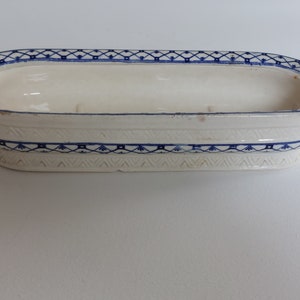 May include: An oval-shaped, off-white ceramic soap dish with blue geometric patterns. The dish has a raised rim and a decorative border of blue floral and geometric designs. The overall design is simple and elegant.