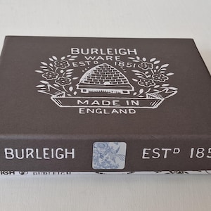 May include: A rectangular brown box with white text and a floral design. The box reads "Burleigh Ware Estd 1851" and "Made in England". A small, square sticker with a floral pattern is on the front.