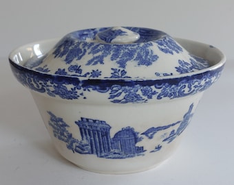 May include: A white and blue ceramic bowl with a lid. The bowl features a blue floral pattern around the rim and a scenic design of buildings. The lid has a central knob and a matching blue floral design. A classic kitchenware item.