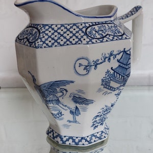 May include: White and blue ceramic pitcher with a hexagonal body. The pitcher features a blue floral and pagoda design. The handle and rim are also trimmed in blue. The pitcher is a decorative piece.
