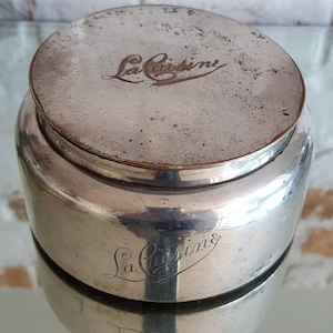 May include: A vintage silver-toned jar with a lid, featuring the engraved text "La Cuisine" in cursive. The jar has a cylindrical body and a slightly domed lid. The surface shows signs of age and wear.