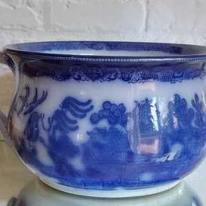 May include: A blue and white ceramic chamber pot with a handle. The pot features a Willow pattern design with a landscape scene. The rim and handle are white, while the body is blue and white.
