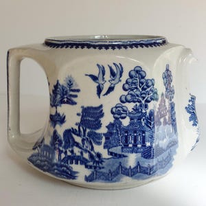 May include: A white ceramic teapot with a blue Willow pattern design. The teapot features a rectangular shape with a handle and spout. The blue design depicts a landscape with trees, buildings, and birds. The teapot is a classic piece of tableware.