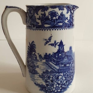 May include: A white ceramic pitcher with a blue and white floral and landscape design. The pitcher features a handle and a spout. The design includes flowers, trees, buildings, and birds. The pitcher is likely vintage.