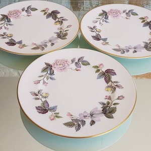 May include: Three white ceramic plates with a floral rose design in pink, purple, and green. The plates have a gold rim and are arranged on a light blue surface. The floral pattern encircles the plate's edge.