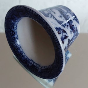 May include: A blue and white ceramic container with a flared rim. The container features a detailed pattern of figures and landscapes. The rim has a decorative border. The interior is white, and the exterior is blue and white.