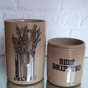 May include: Two beige ceramic kitchen storage jars. The larger jar has a black illustration of celery and the word "CELERY" at the bottom. The smaller jar has the words "BEEF DRIPPING" in black lettering.
