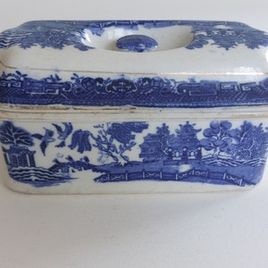 May include: Rectangular blue and white ceramic dish with lid. The dish features a Willow pattern design with a landscape scene. The lid has a small knob and the edges are trimmed with gold. A classic piece for serving or display.