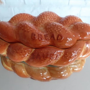 May include: A ceramic bread and sauce dish in the shape of a braided loaf. The dish is glazed in shades of brown and tan, with the word "BREAD" embossed on the lid and "SAUCE" on the base. The dish has a glossy finish.