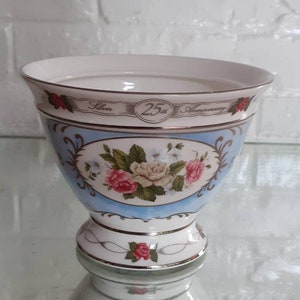 May include: A decorative ceramic bowl with a light blue exterior, featuring floral designs in pink, white, and green. The bowl has a gold trim and the text "Silver 25th Anniversary". The bowl is on a pedestal base.