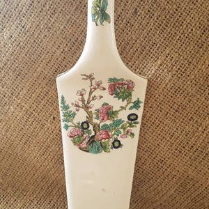 Puede incluir: Un servidor de pastel de cerámica color crema con un diseño floral. El servidor tiene un mango largo y una hoja ancha. El diseño floral incluye rosas rosas y rojas, hojas verdes y pequeñas flores azules. Mide unos 28 cm de largo.