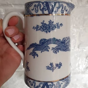 Puede incluir: Jarra de cerámica blanca con un diseño de dragón y flores azules. La jarra tiene un borde y un asa dorados. El diseño es de estilo clásico azul y blanco, con una estética asiática tradicional.