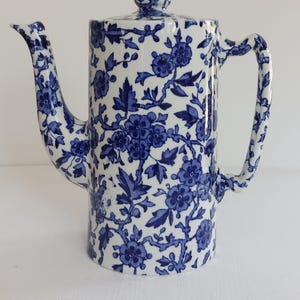 May include: A white and blue floral patterned ceramic coffee pot. The pot has a tall, cylindrical shape with a curved spout and handle. The lid is topped with a small, round knob. The blue floral design covers the entire surface of the pot.