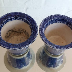 May include: Two blue and white ceramic vases with a detailed pattern around the rim. The vases have a classic design, with a light interior and a dark blue exterior. The vases are reflected on a shiny surface.