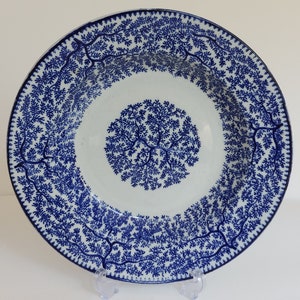 May include: A white ceramic serving bowl with a blue tree branch pattern. The bowl has a wide rim and a central design of a tree branch pattern. The bowl is displayed on a clear plastic stand.