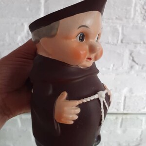 May include: A vintage ceramic figurine of a monk, featuring a brown robe and hat, with a white rope belt. The figurine has a detailed face with rosy cheeks and a slightly stern expression. The base is black.