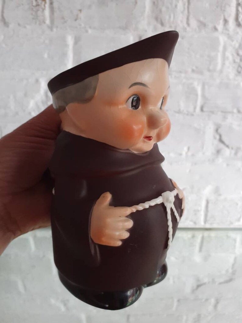 May include: A vintage ceramic figurine of a monk, featuring a brown robe and hat, with a white rope belt. The figurine has a detailed face with rosy cheeks and a slightly stern expression. The base is black.