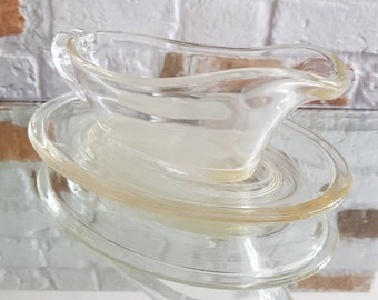 May include: A clear glass gravy boat with a matching saucer. The gravy boat has a curved handle and a pouring spout. The saucer has concentric rings. The set is made of clear glass.