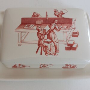 May include: A rectangular, off-white ceramic butter dish with a lid. The lid features a red illustration of people working at a table. The base of the dish is also off-white.