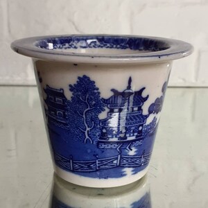 May include: A small, white and blue ceramic planter with a classic Willow pattern design. The planter features a detailed landscape scene with pagodas, trees, and a fence. The rim of the planter is slightly flared.