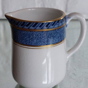 May include: A white ceramic creamer with a blue and gold patterned band around the middle. The creamer has a handle and a spout for easy pouring. The design includes a dotted pattern and a gold trim.