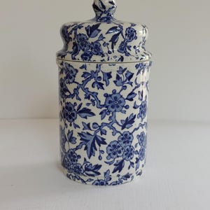 May include: A ceramic jar with a lid, featuring a blue floral pattern on a white background. The jar has a cylindrical shape with a slightly wider base and a matching lid with a small knob. The floral design includes flowers and leaves.