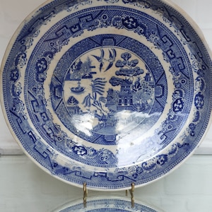 May include: A blue and white ceramic plate with a detailed Willow pattern design. The plate features a central landscape scene with pagodas, trees, and a boat, surrounded by intricate borders. The plate is displayed on a gold-colored stand.